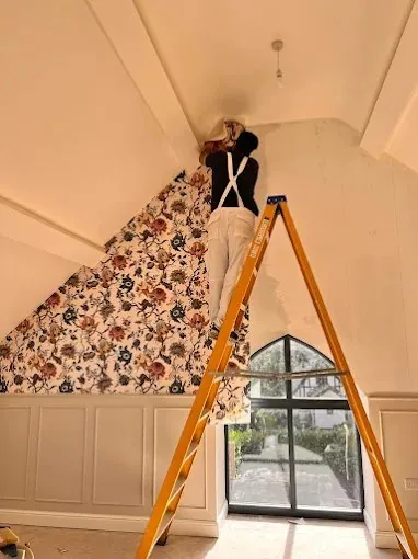 A decorator hanging bold floral wallpaper up a sloped ceiling